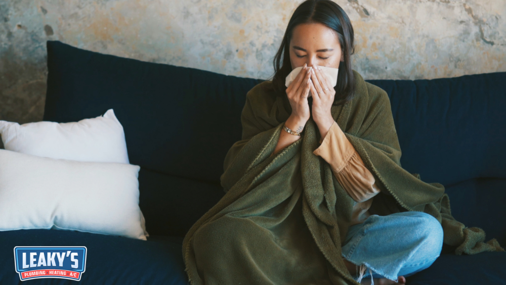 Woman sitting indoors wrapped in a blanket and sneezing into a tissue, representing allergy symptoms and poor indoor air quality.