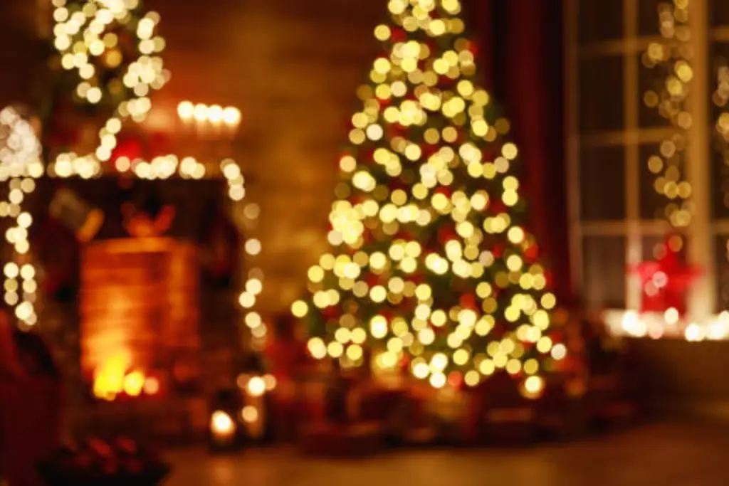 Decorated Christmas tree with glowing lights and ornaments beside a fireplace with stockings, garlands, and wrapped presents on the floor in a warmly lit living room.