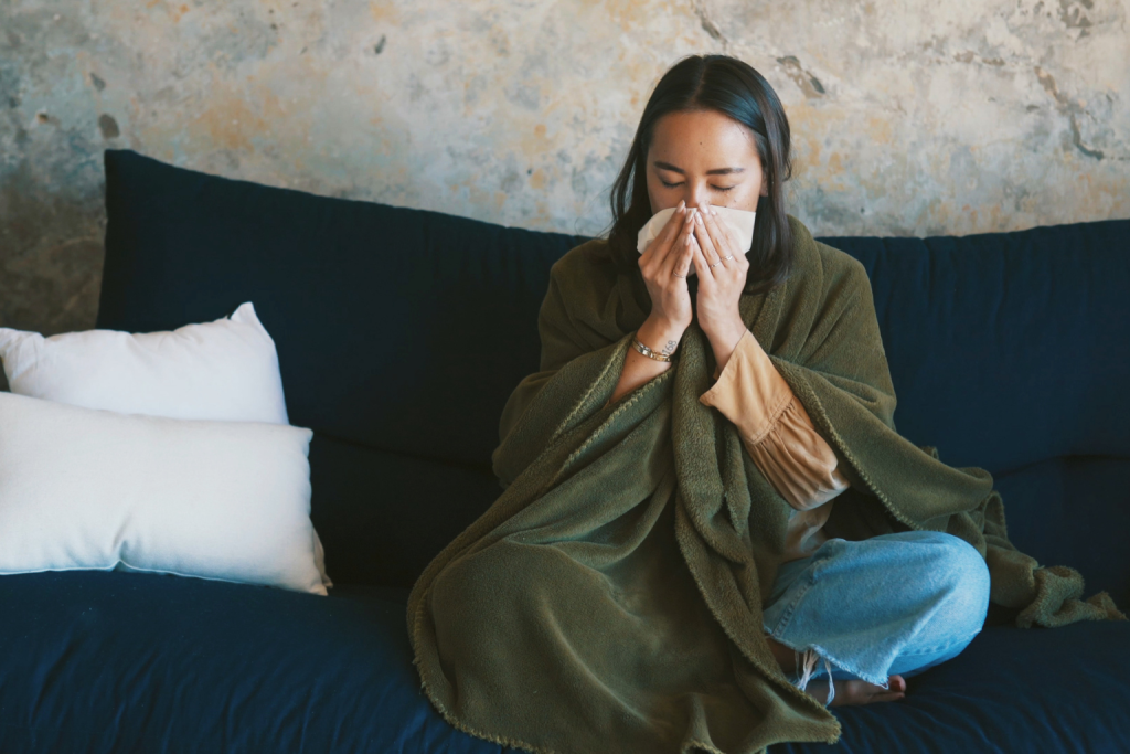 Woman sitting indoors wrapped in a blanket and sneezing into a tissue, representing allergy symptoms and poor indoor air quality.