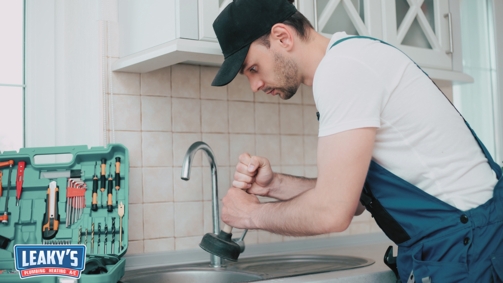 A professional plumber unclogging a kitchen sink with a plunger, with a toolkit of repair tools placed beside the sink in a modern home kitchen.